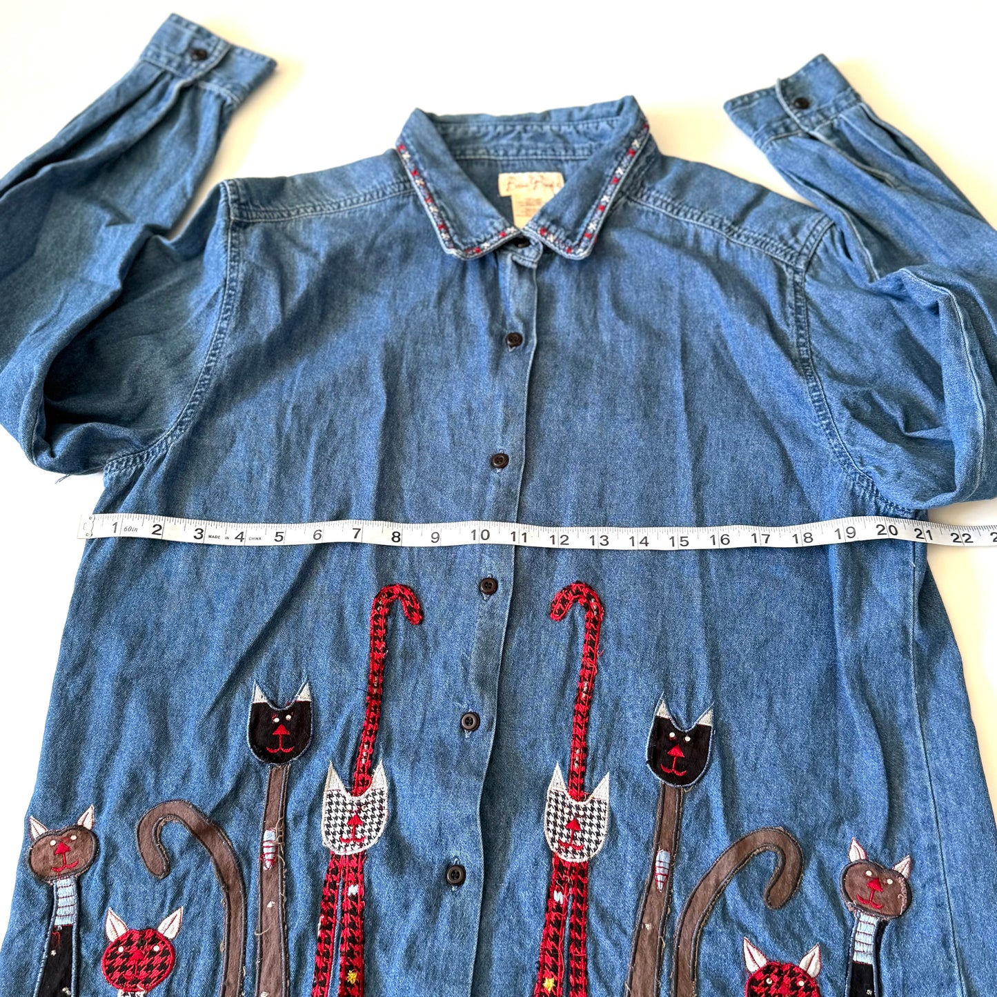 Blue denim shirt with embroidered cat designs and a measuring tape for size reference.