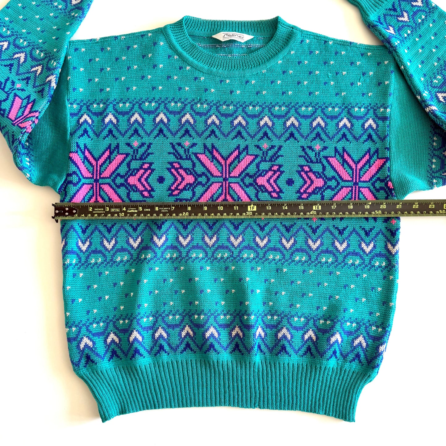 Teal and pink sweater with an aztec / snowflake adjacent style pattern, on a white background and a measuring tape laid on it.