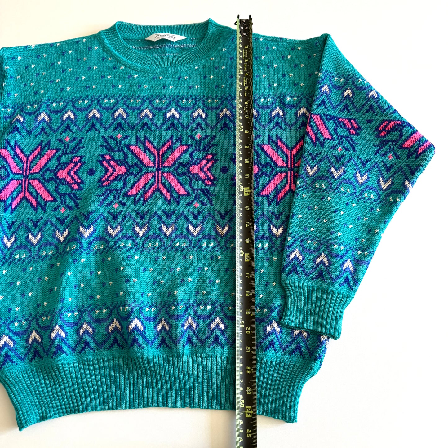 Teal and pink sweater with an aztec / snowflake adjacent style pattern, on a white background and a measuring tape laid on it. 