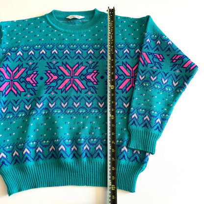 Teal and pink sweater with an aztec / snowflake adjacent style pattern, on a white background and a measuring tape laid on it. 