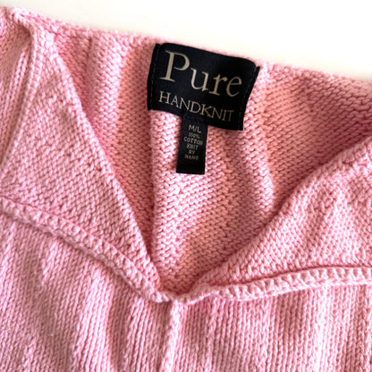 Close up of the tag reading "PURE HANDKNIT M/L 100% COTTON KNIT BY HAND" of the pink tank top on white backdrop.