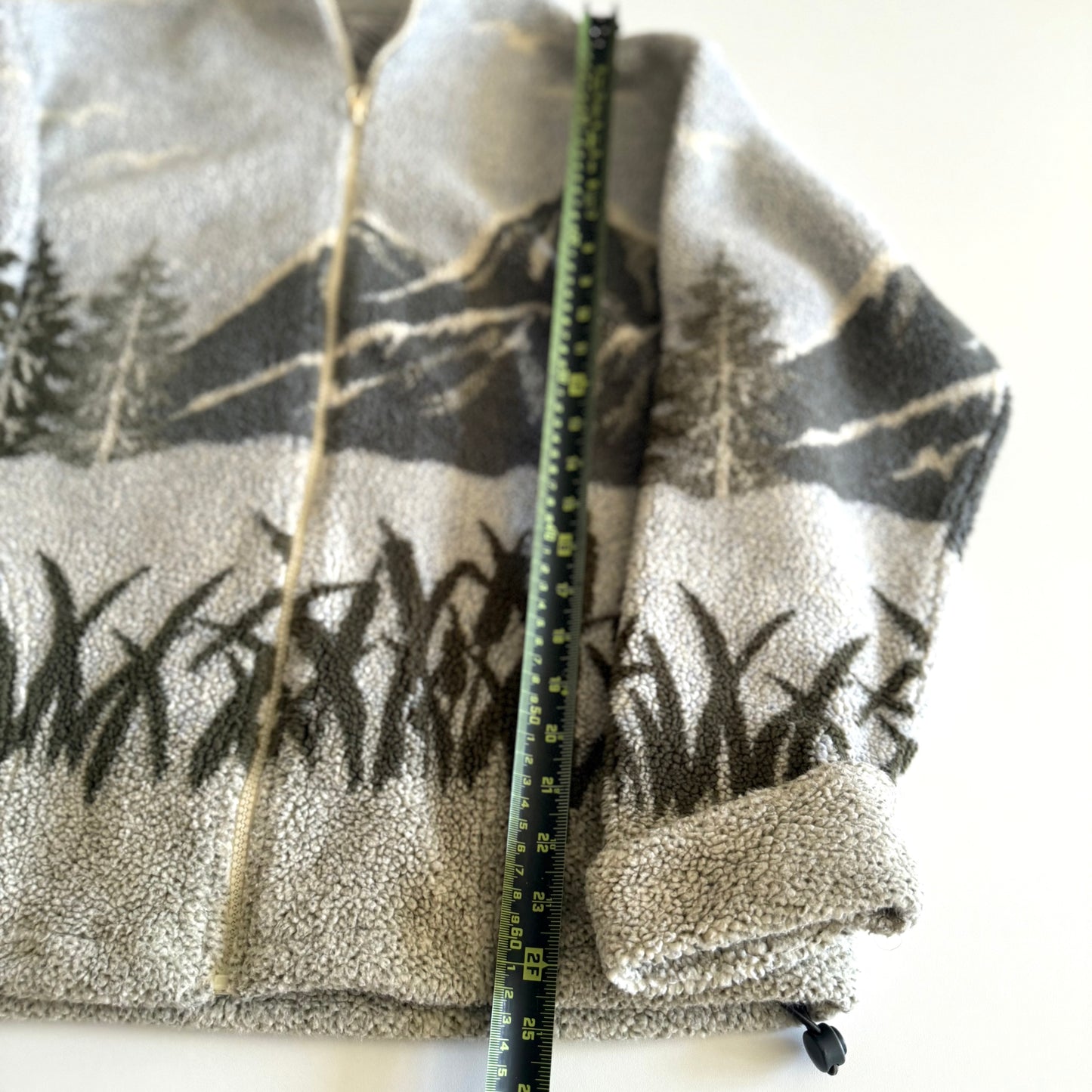 Fleece textured sweater with a mountain marsh forest scene on it, and a zippered close - on a white backdrop with a measuring tape laid  on it. 