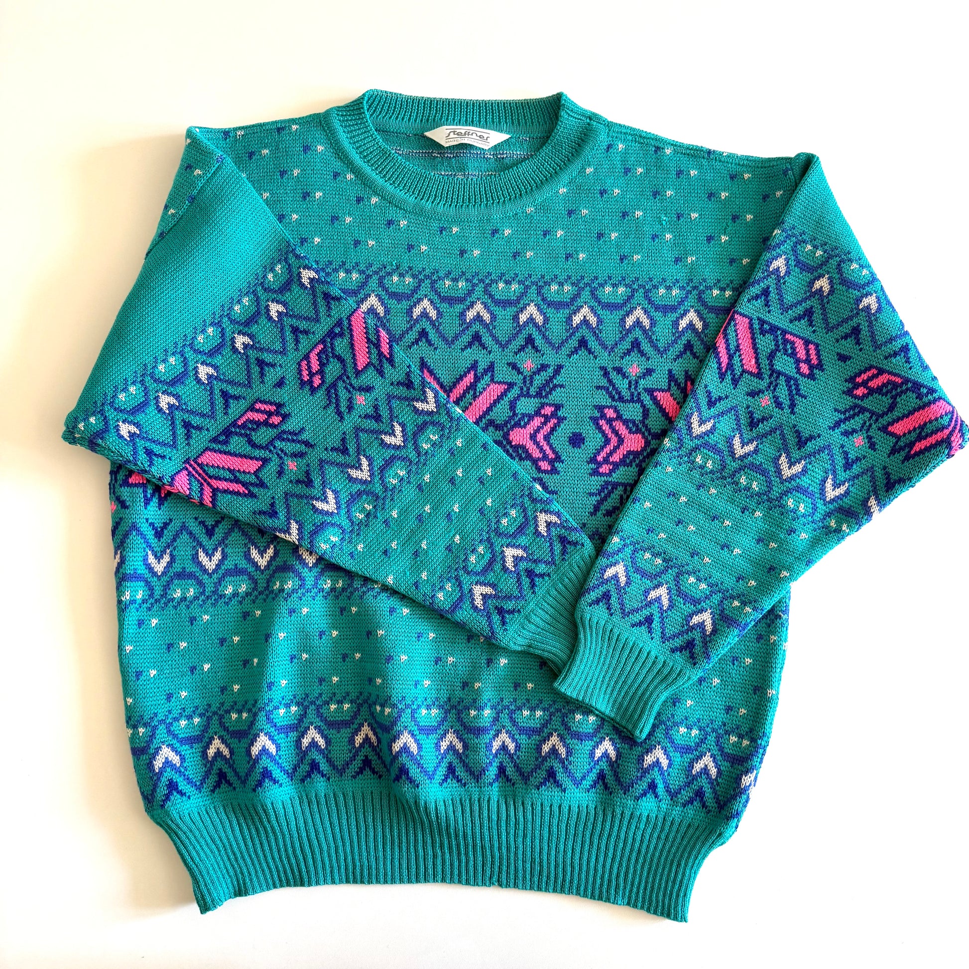 Teal and pink sweater with an aztec / snowflake adjacent style pattern, on a white background. 