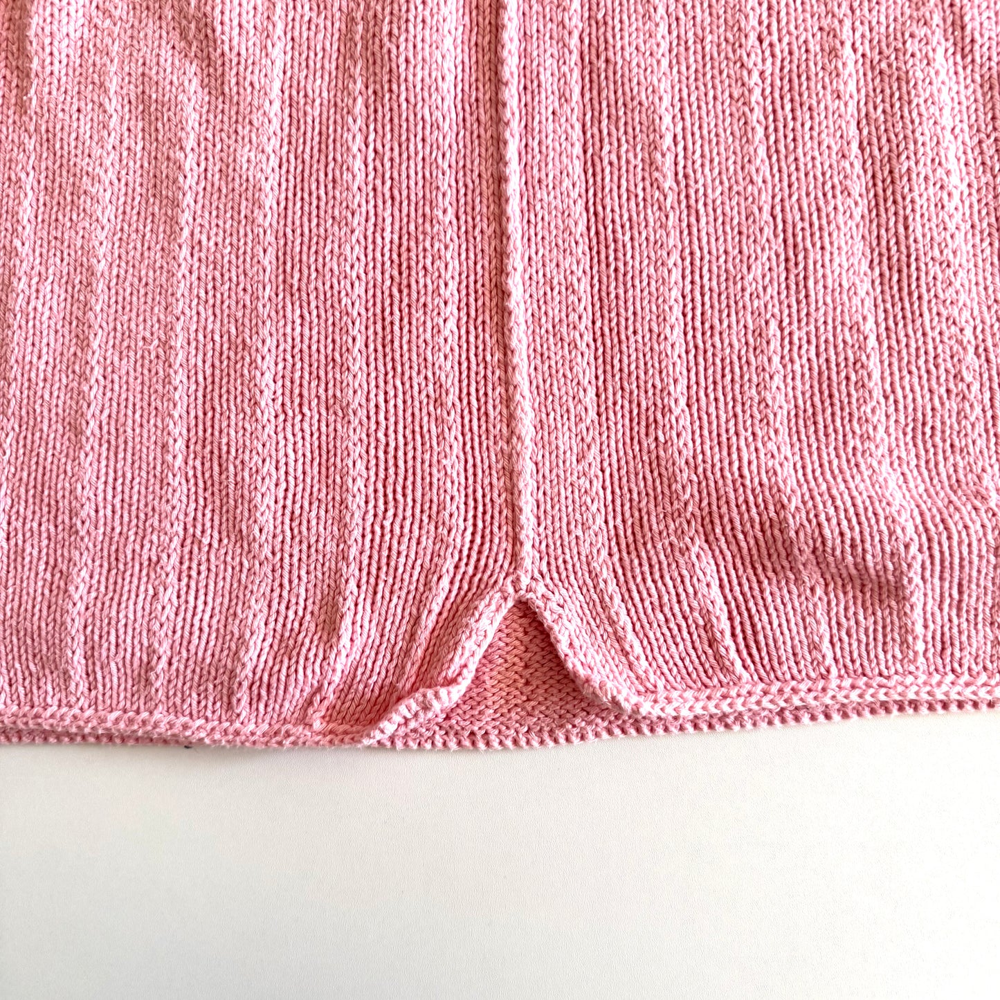 Close up of the bottom of the pink knit tank top, of a small (manufactured) split at the base of the centre of the shirt - on a white backdrop. 
