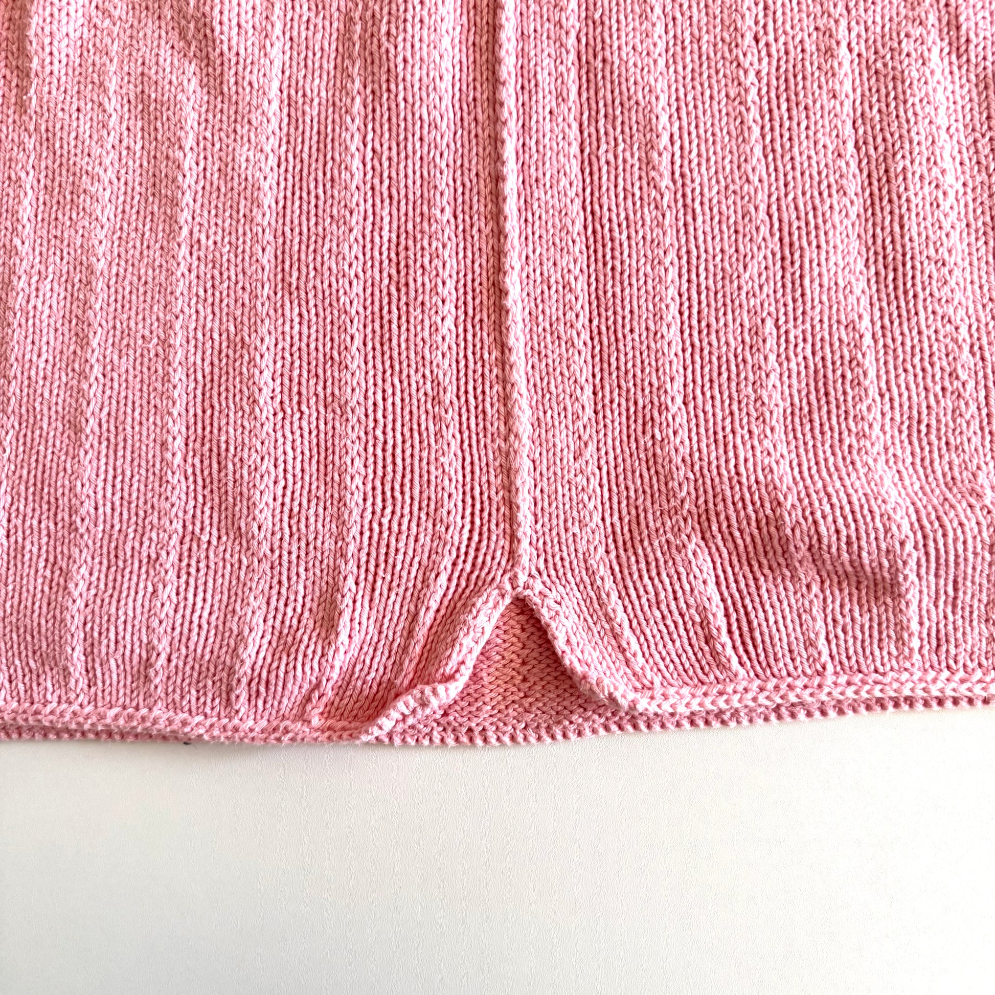 Close up of the bottom of the pink knit tank top, of a small (manufactured) split at the base of the centre of the shirt - on a white backdrop. 