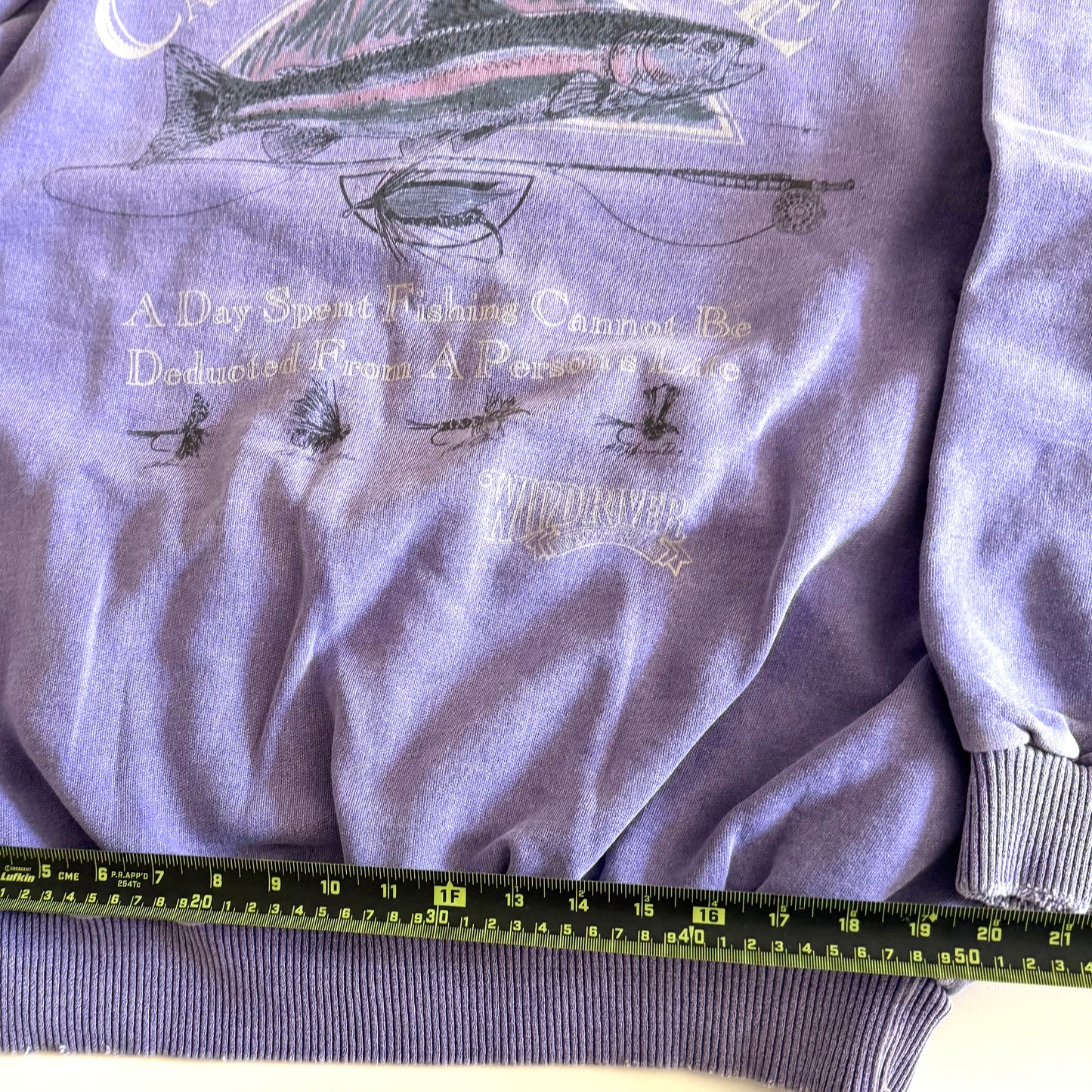 Light purple sweatshirt with fishing theme images and words on the front, on a white backdrop - with a measuring tape laid on it. 