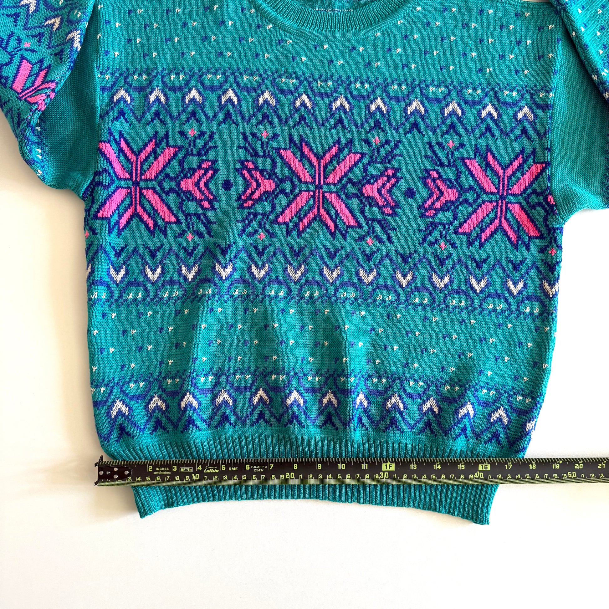 Teal and pink sweater with an aztec / snowflake adjacent style pattern, on a white background and a measuring tape laid on it. 
