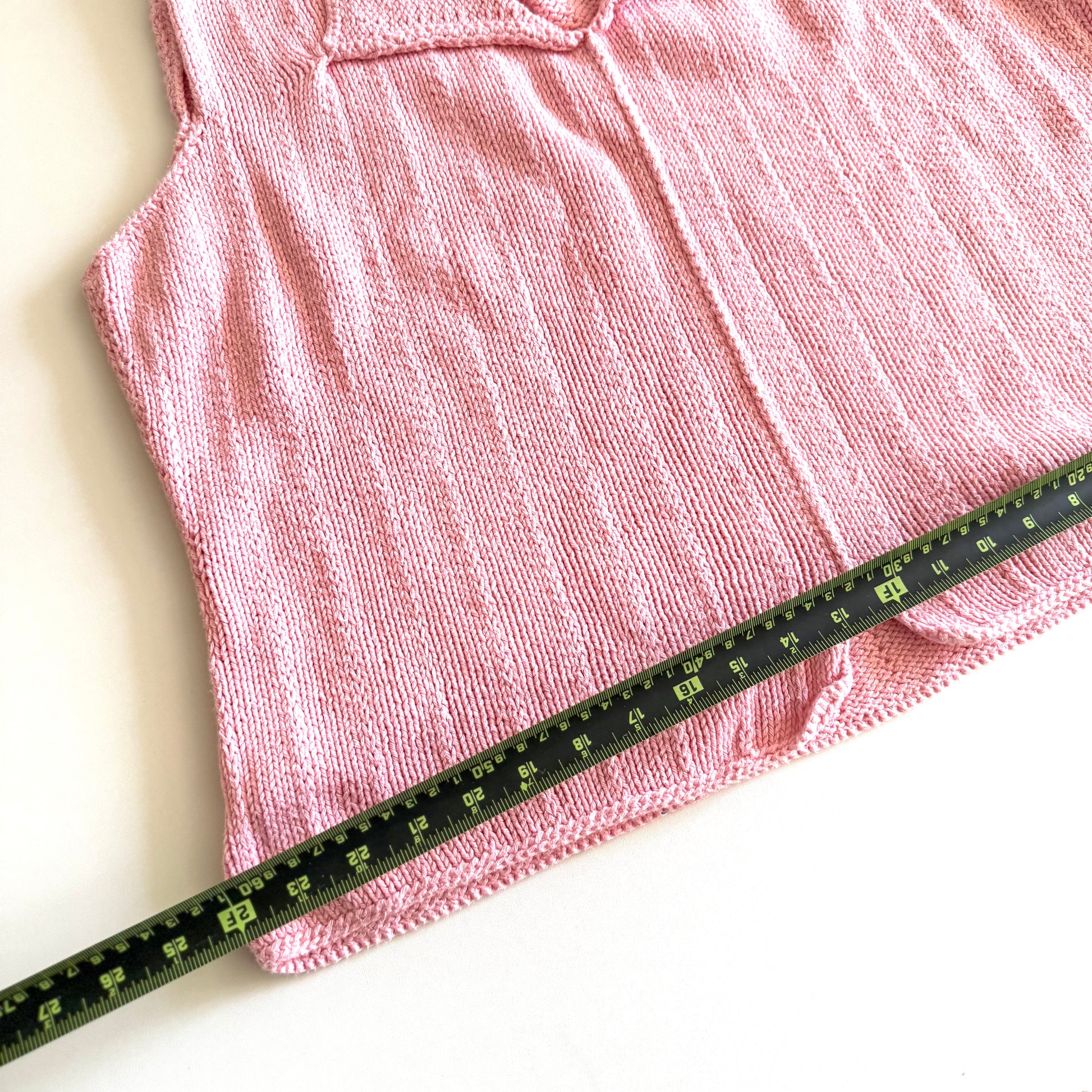 Light pink knit style tank top, with folded out collar opening on a white backdrop - with a measuring tape laid on top. 