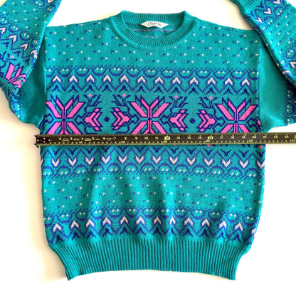 Teal and pink sweater with an aztec / snowflake adjacent style pattern, on a white background and a measuring tape laid on it. 