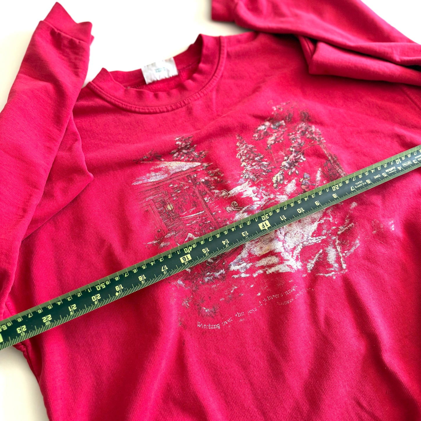 Bold pink sweatshirt / crewneck with a winter scene on the front  laid on a white backdrop - with a measuring tape laid out on it. 