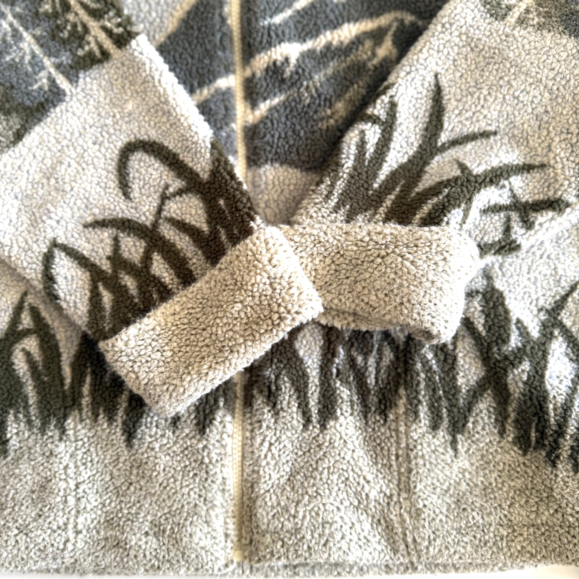 Fleece textured sweater with a mountain marsh forest scene on it, and a zippered close - on a white backdrop.  Close up of the sleeves. 