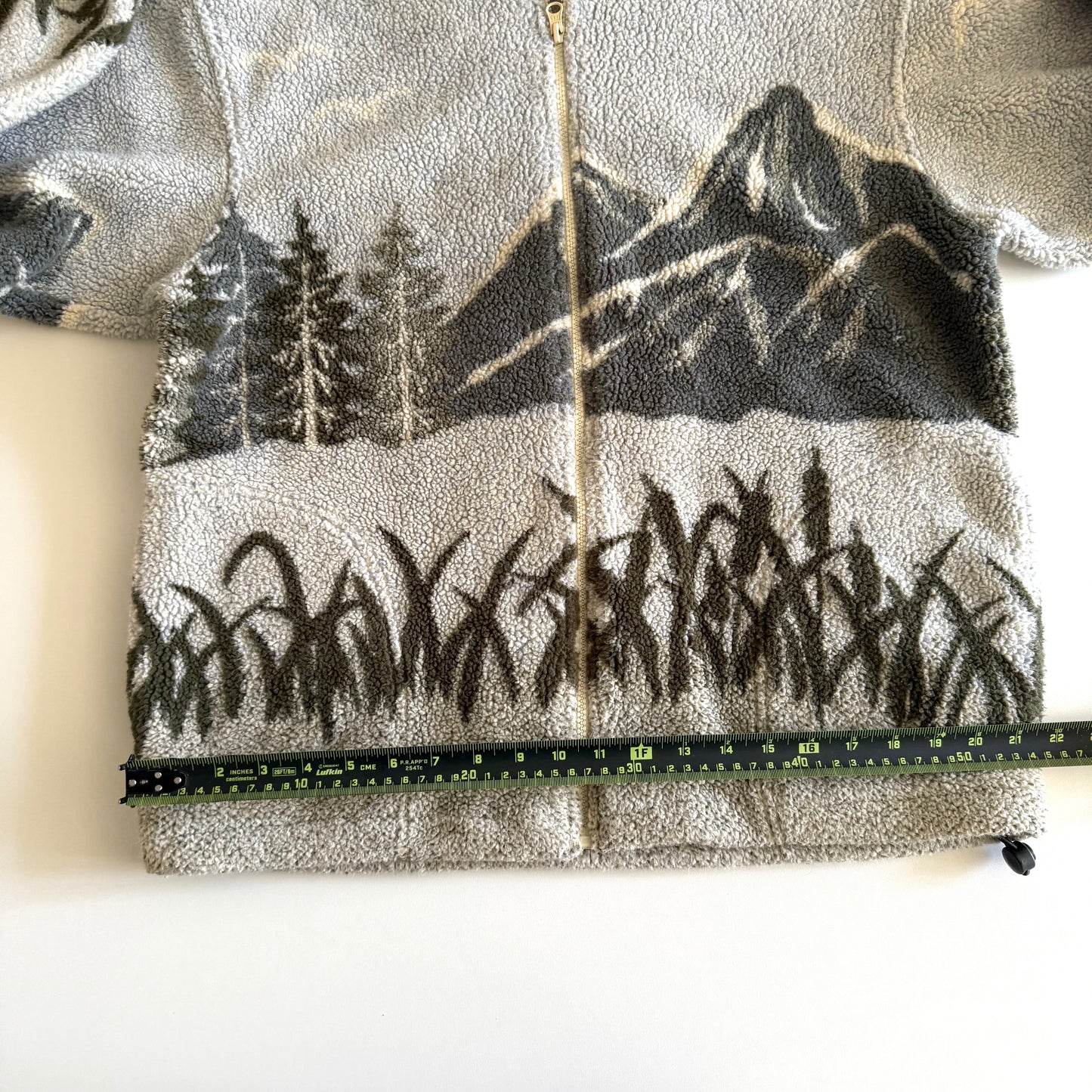 Fleece textured sweater with a mountain marsh forest scene on it, and a zippered close - on a white backdrop with a measuring tape laid  on it. 