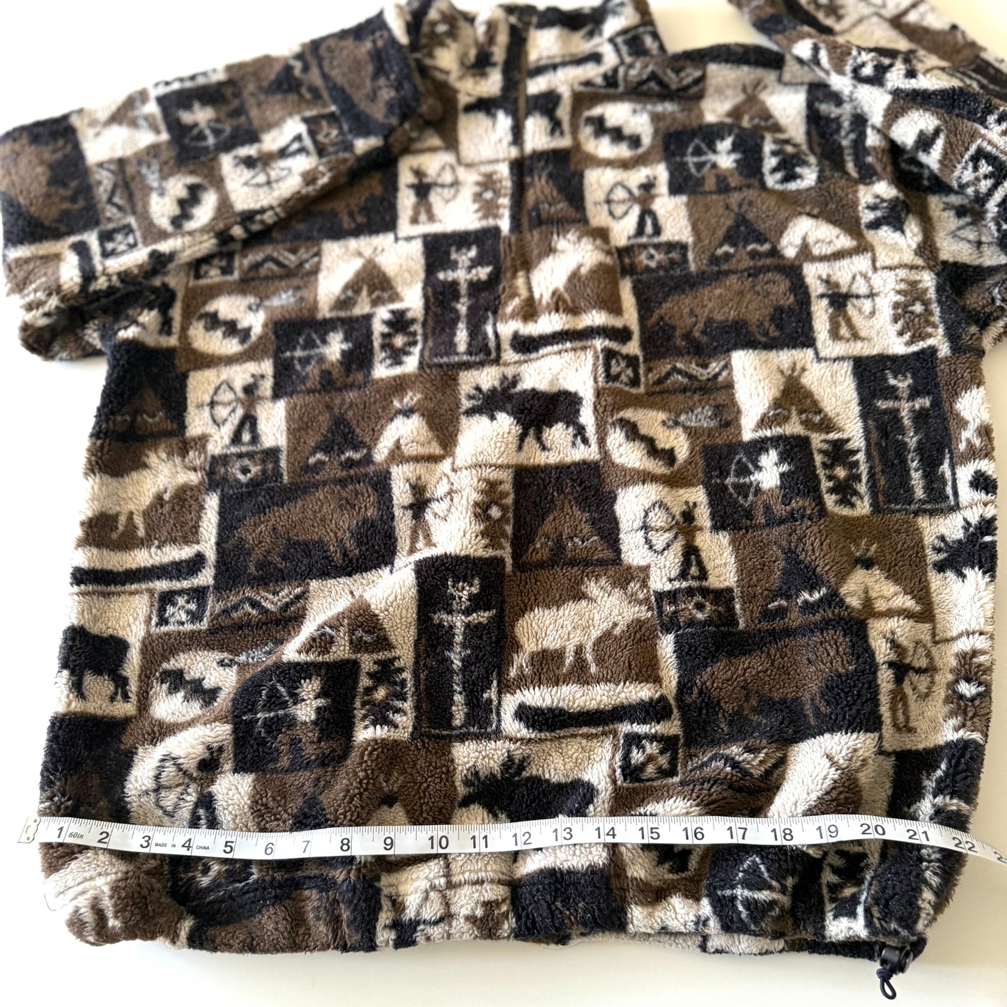 Moose and other camping motif quarter zip fleece on a white background with a measuring tape on it. 