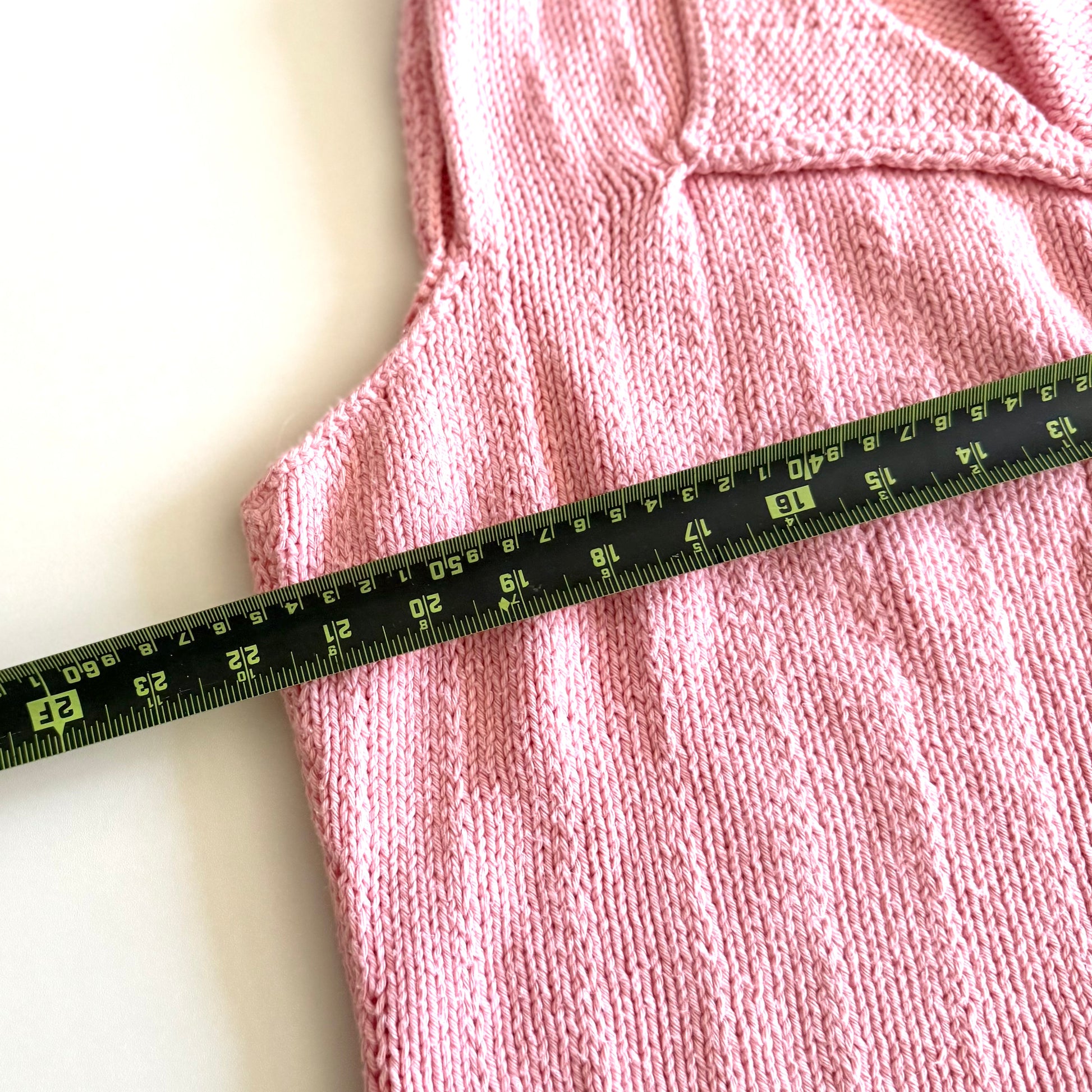 Light pink knit style tank top, with folded out collar opening on a white backdrop - with a measuring tape laid on top. 