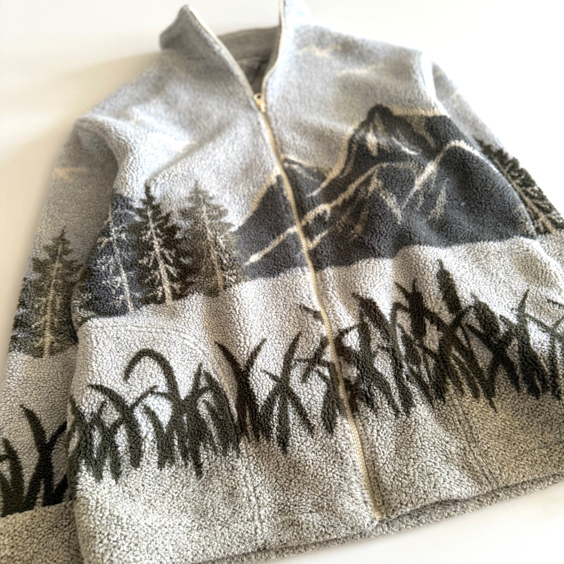 Fleece textured sweater with a mountain marsh forest scene on it, and a zippered close - on a white backdrop. 