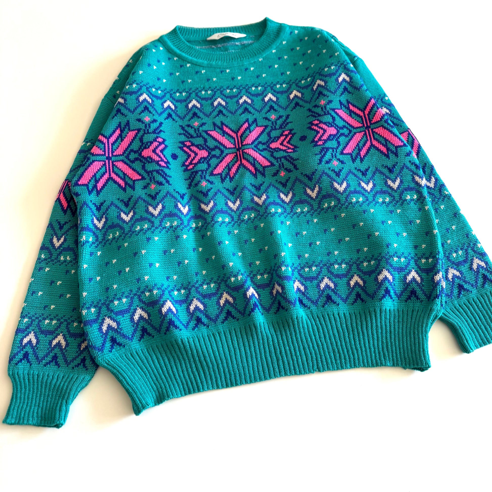 Teal and pink sweater with an aztec / snowflake adjacent style pattern, on a white background. 