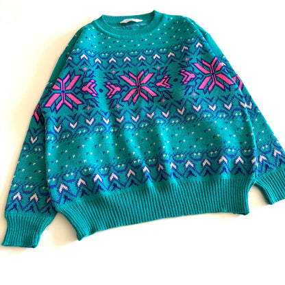Teal and pink sweater with an aztec / snowflake adjacent style pattern, on a white background. 