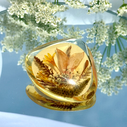 Transparent resin brooch with pressed flowers on a reflective surface with white flowers in the background