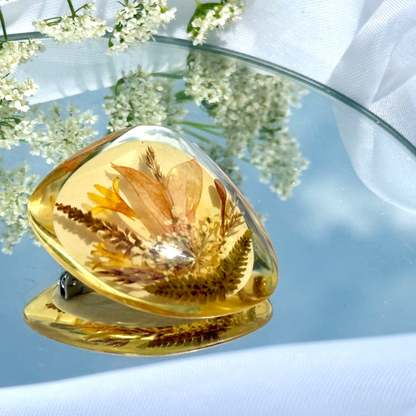 Amber-colored brooch with pressed flowers on a reflective surface with white flowers in the background