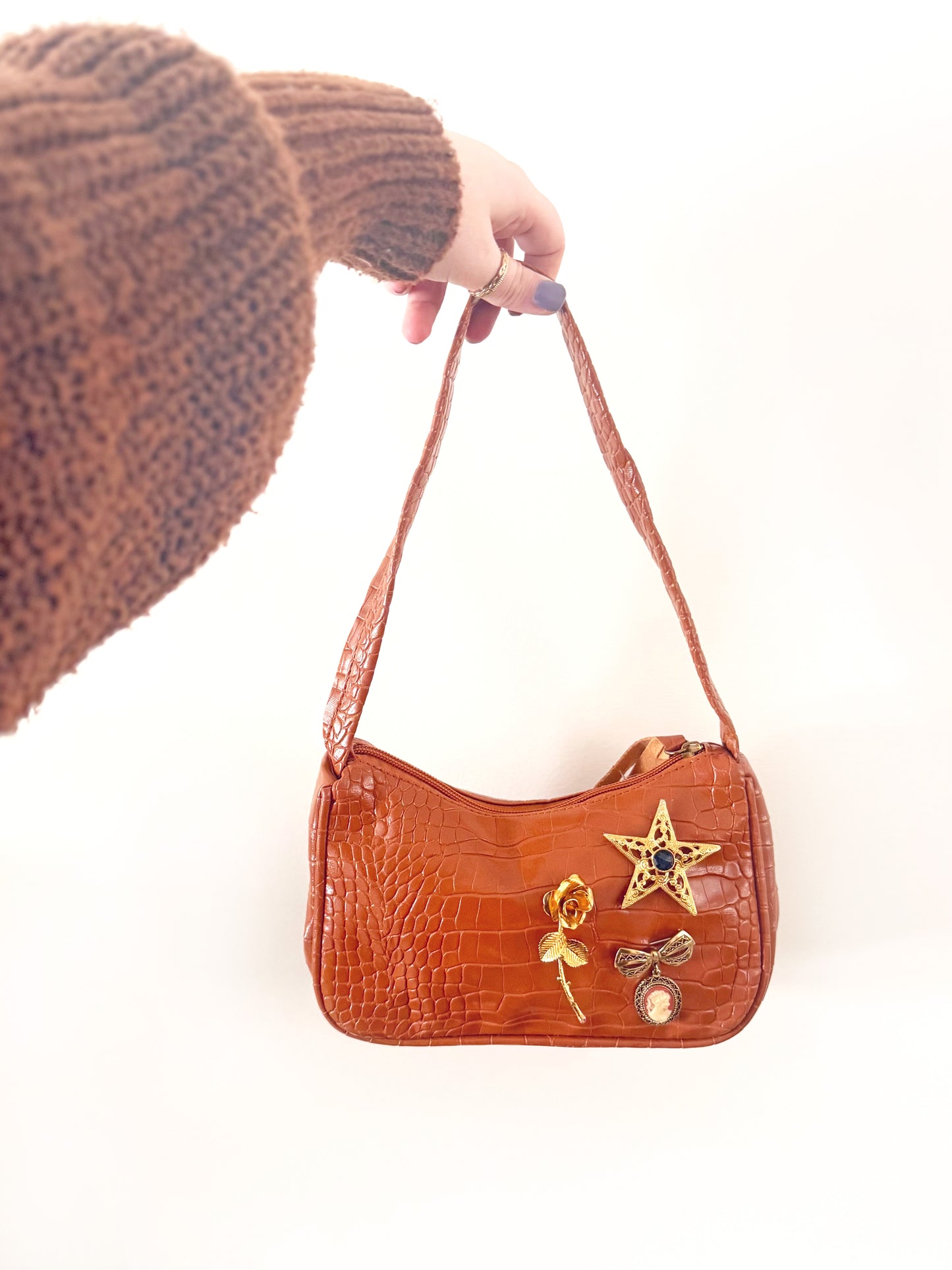 Brown handbag with decorative elements held by a person wearing a brown knitted hat.