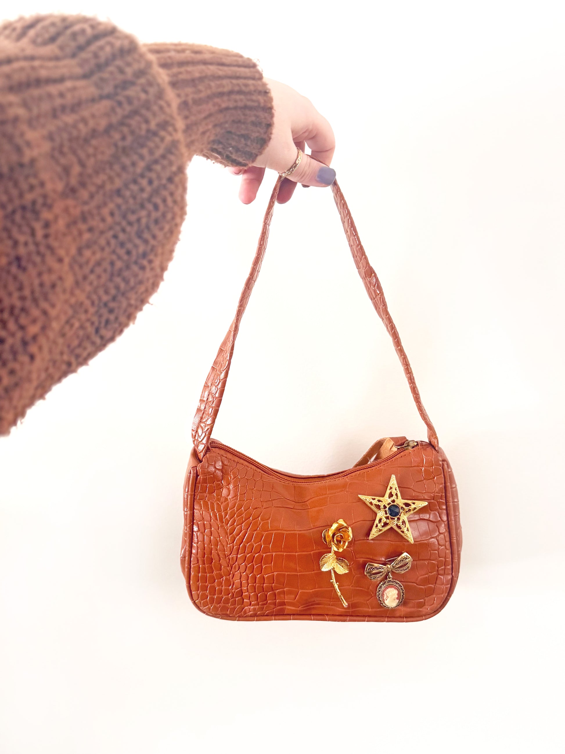 Brown handbag with decorative elements held by a person wearing a brown knitted hat.